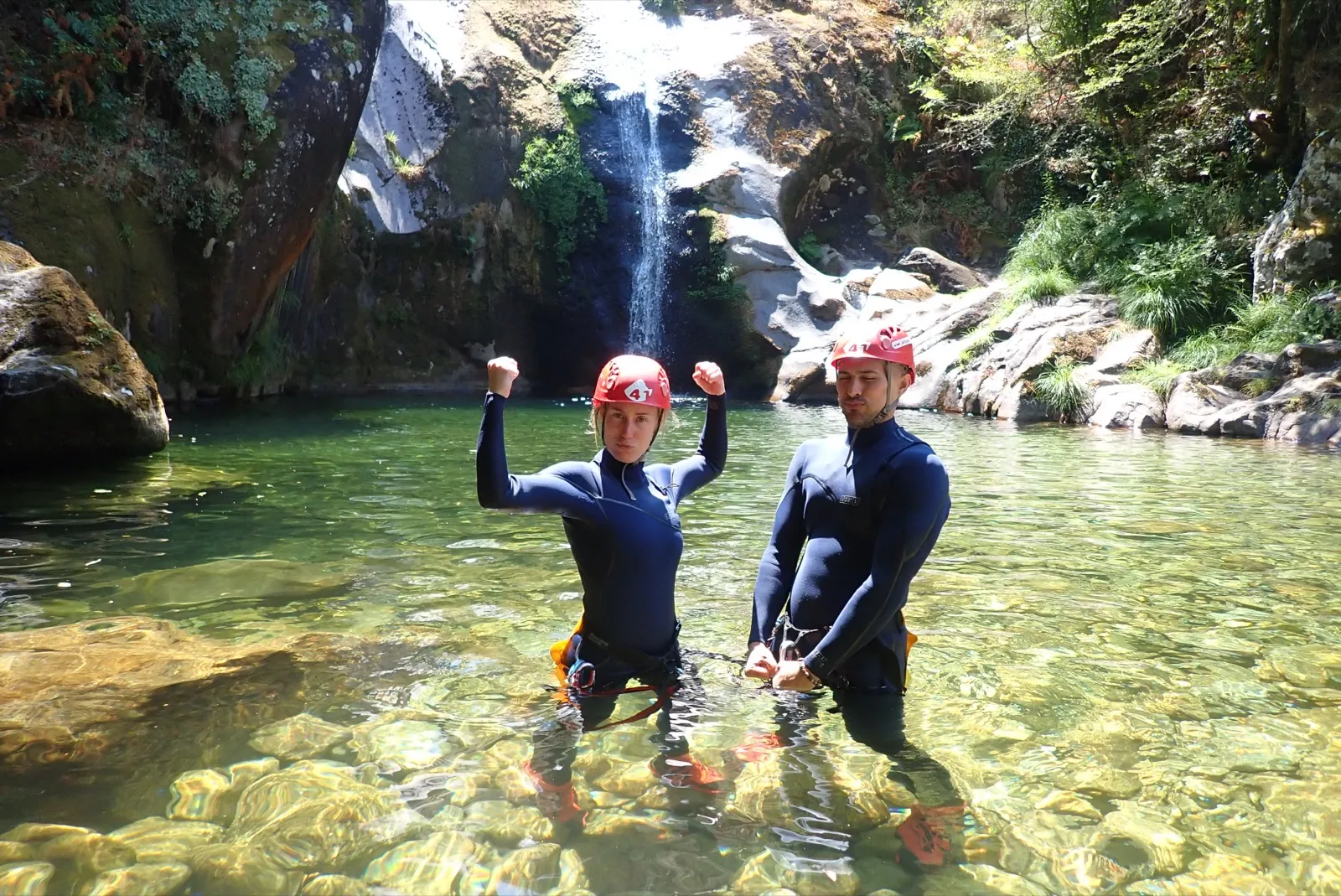 Anne and David flexing in wetsuits by a waterfall.